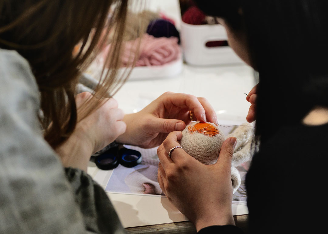 Two people are focused on painting an orange pattern on a white fabric ball, with yarn and scissors on the table.