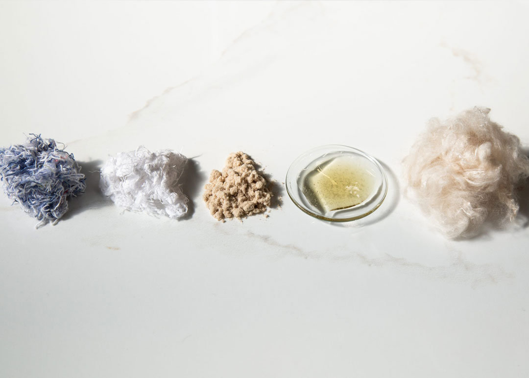 A variety of textile materials displayed on a white surface, including a pile of blue and white fibers on the left, a white fluffy material next to it, a mound of beige fibers in the center, a small glass dish containing a yellowish liquid, and a fluffy white substance on the far right.